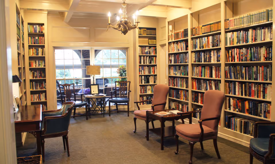 Cozy library-style common room with bookshelves lining the walls, upholstered chairs, small tables, and a chandelier.