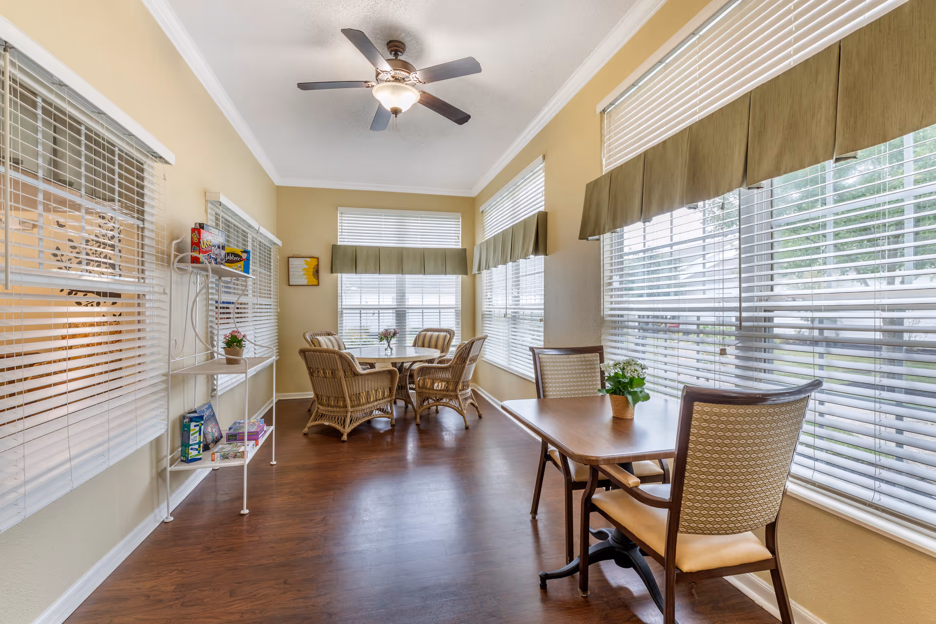A bright room with large windows covered by white blinds and green valances. The room has wooden flooring and a ceiling fan with a light. There are two seating areas: one with a round table surrounded by four wicker chairs, and another with a rectangular wooden table and two cushioned chairs. A white metal shelf on the left holds board games and a small potted plant. The walls are painted light beige.