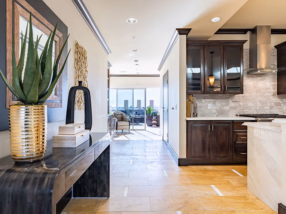 View down a hallway in a senior living facility showing a modern kitchen with dark wood cabinets and stainless steel range hood on the right, and a decorative console table with a potted plant and decorative items on the left. At the end of the hallway, there is a seating area with chairs and large windows letting in natural light.