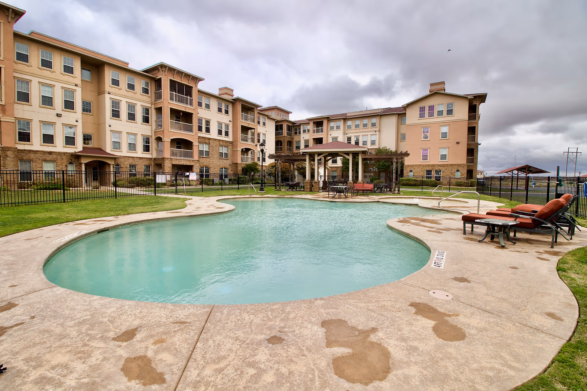 Outdoor swimming pool area with lounge chairs and tables under a gazebo, surrounded by a multi-story residential building under a cloudy sky.
