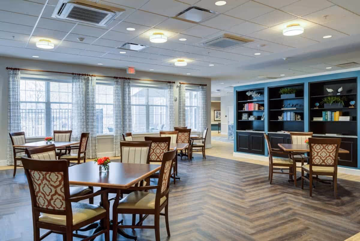 Bright communal dining room with multiple tables and patterned chairs, large windows, and built-in shelving.