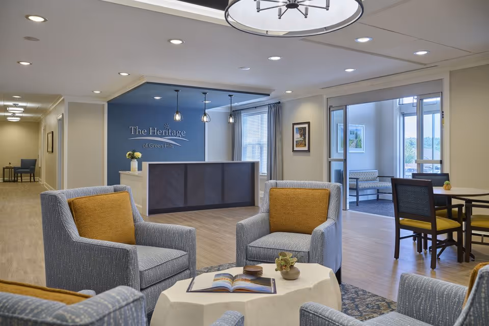 Reception and lounge area with upholstered chairs around a low table, a reception desk, and a wall sign reading 'The Heritage of Green Hills'.