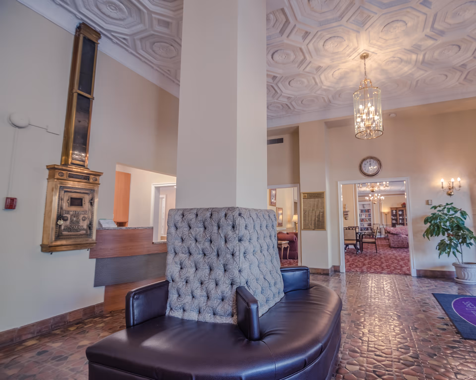 Interior view of a senior living facility lobby area with a central column surrounded by a circular cushioned bench. The ceiling features an ornate geometric pattern with a hanging chandelier. There is a vintage brass mailbox mounted on the wall to the left, a clock above a doorway, and a potted plant on the right. Through the doorway, another room with chairs, tables, and bookshelves is visible.