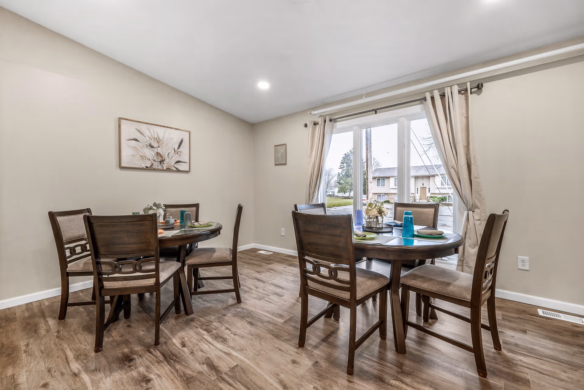 Bright dining room with two round wooden tables and several chairs on a wood floor, a large window with curtains looking out to neighboring houses.
