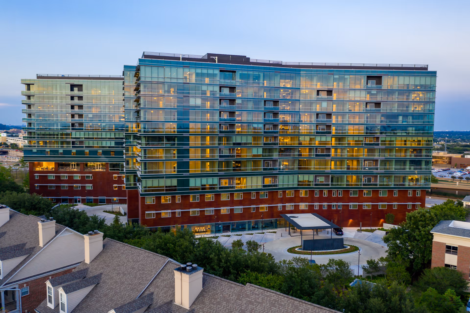 A large modern multi-story building with glass windows and balconies, surrounded by trees and other buildings, taken during dusk with lights visible inside.