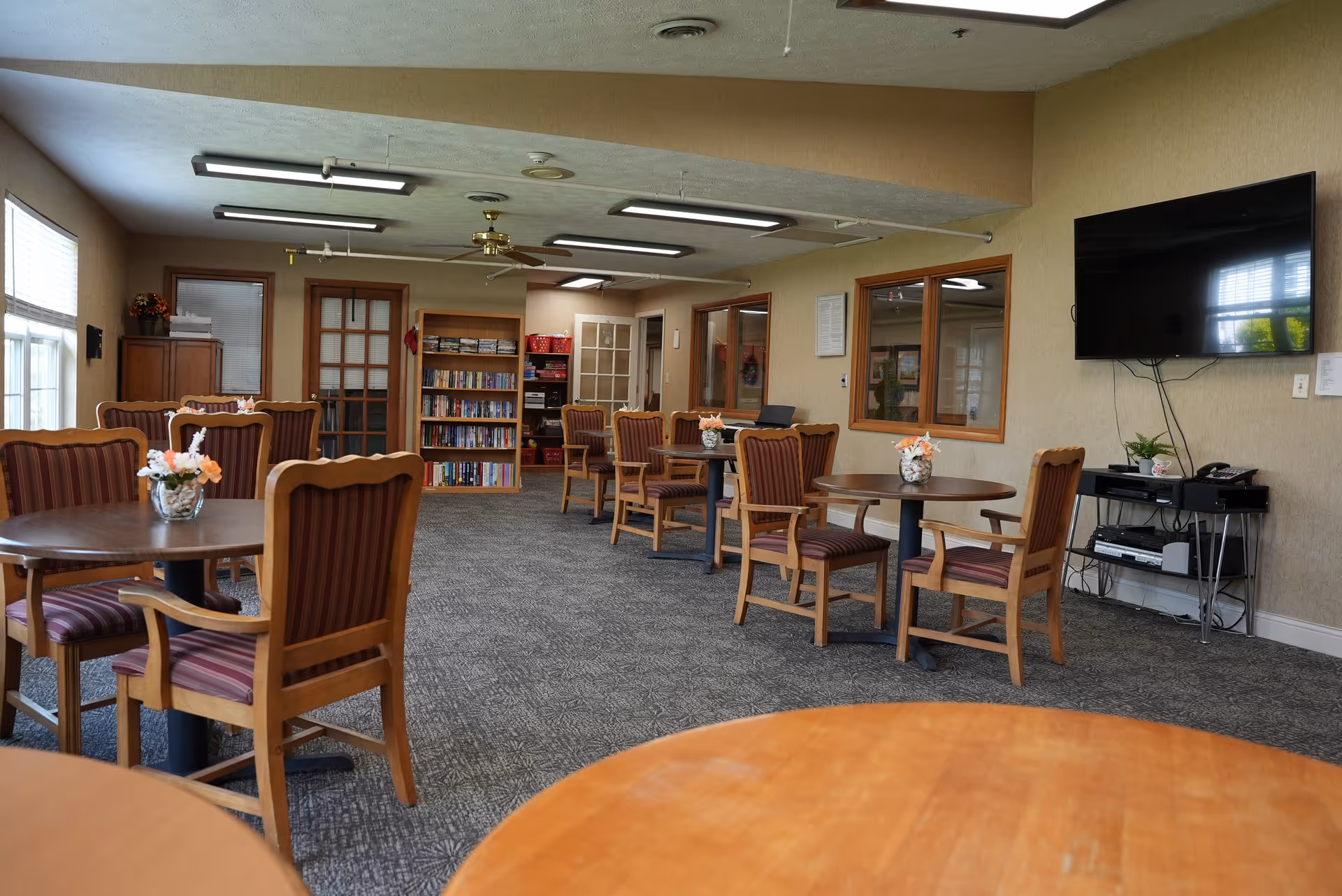 Communal dining/activity room with round tables and wooden chairs, a wall-mounted TV, and bookshelves.