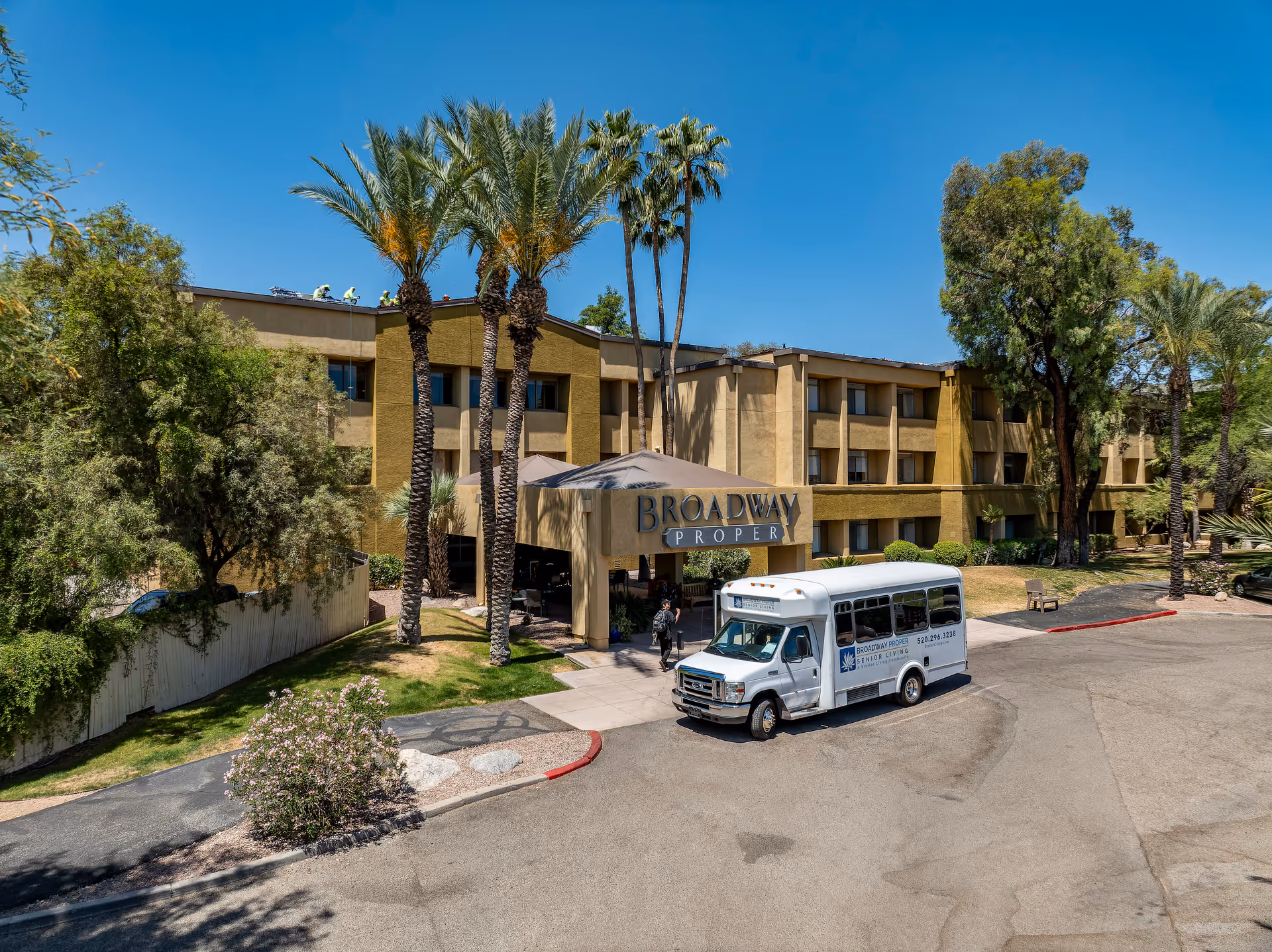 Exterior view of Broadway Proper Assisted & Senior Living facility showing a three-story building with palm trees and other greenery around. A white shuttle bus with the facility's name and phone number is parked near the entrance, where a person is walking towards the building under a covered entryway.