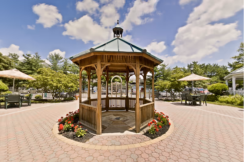 A wooden gazebo with a green roof situated in the center of a paved circular area surrounded by flower beds with red and yellow flowers. There are outdoor tables with umbrellas and chairs around the gazebo, with trees and a clear blue sky with clouds in the background.