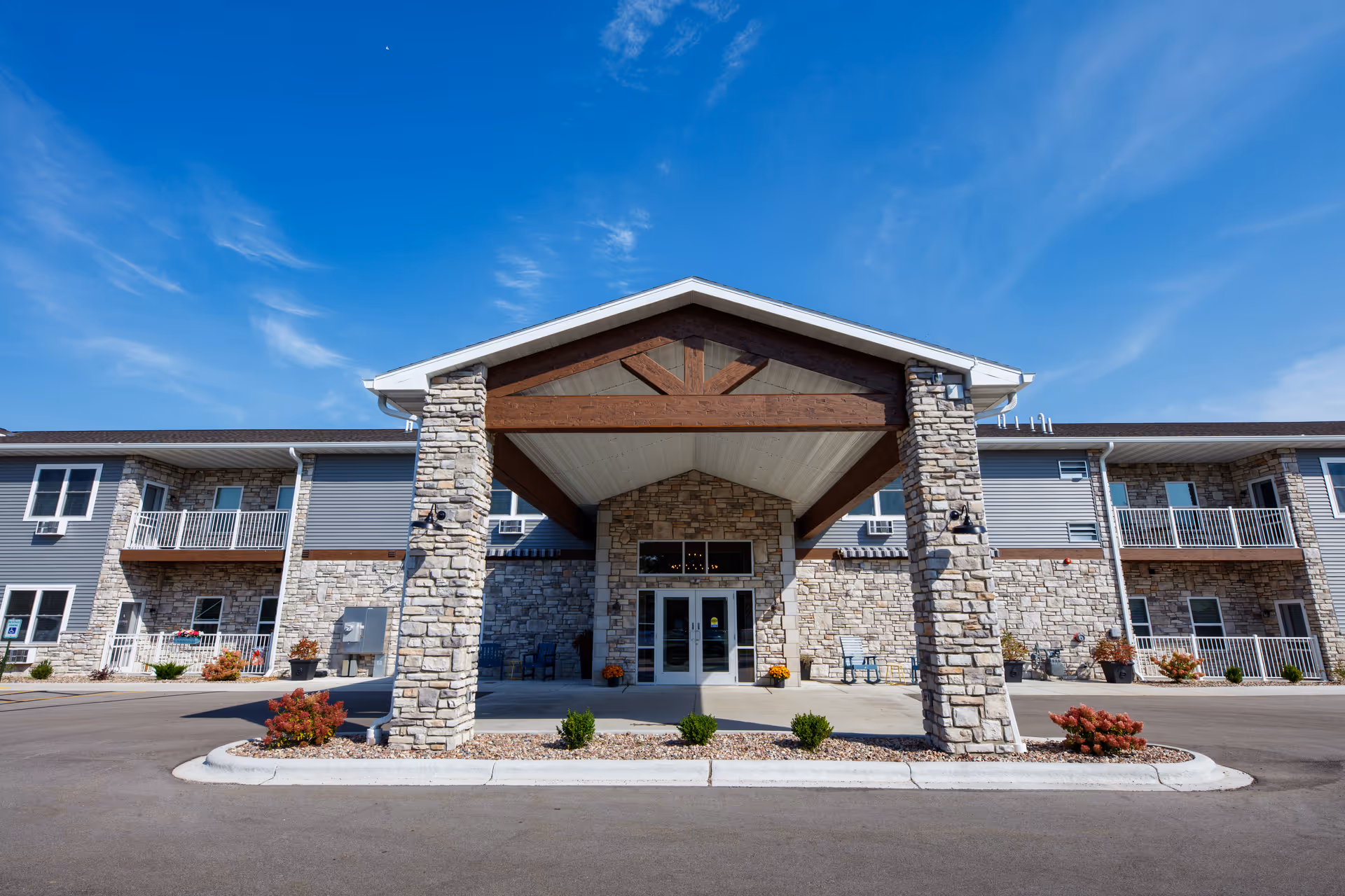 Front exterior view of The Cottages at Lake Park Senior Living & Memory Care building with stone pillars supporting a covered entrance, small shrubs and plants around the entrance, and a clear blue sky above.