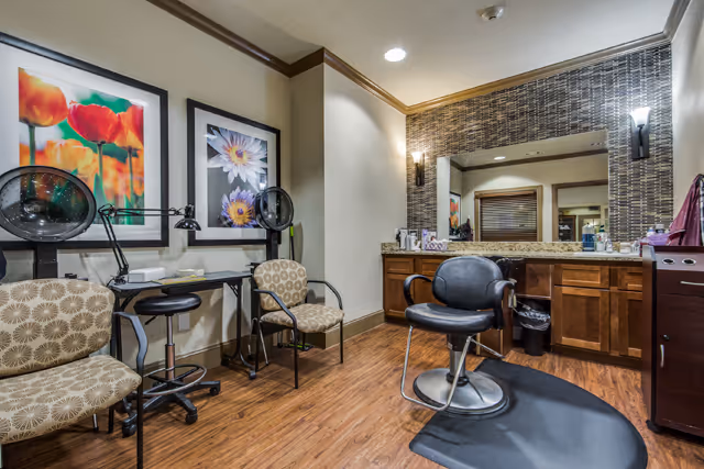 Interior view of a salon or grooming area in a senior living facility with a styling chair on a black mat, two patterned chairs, a small desk with a lamp, and framed floral artwork on the wall. There is a large mirror above a wooden counter with various items and two wall-mounted lights.