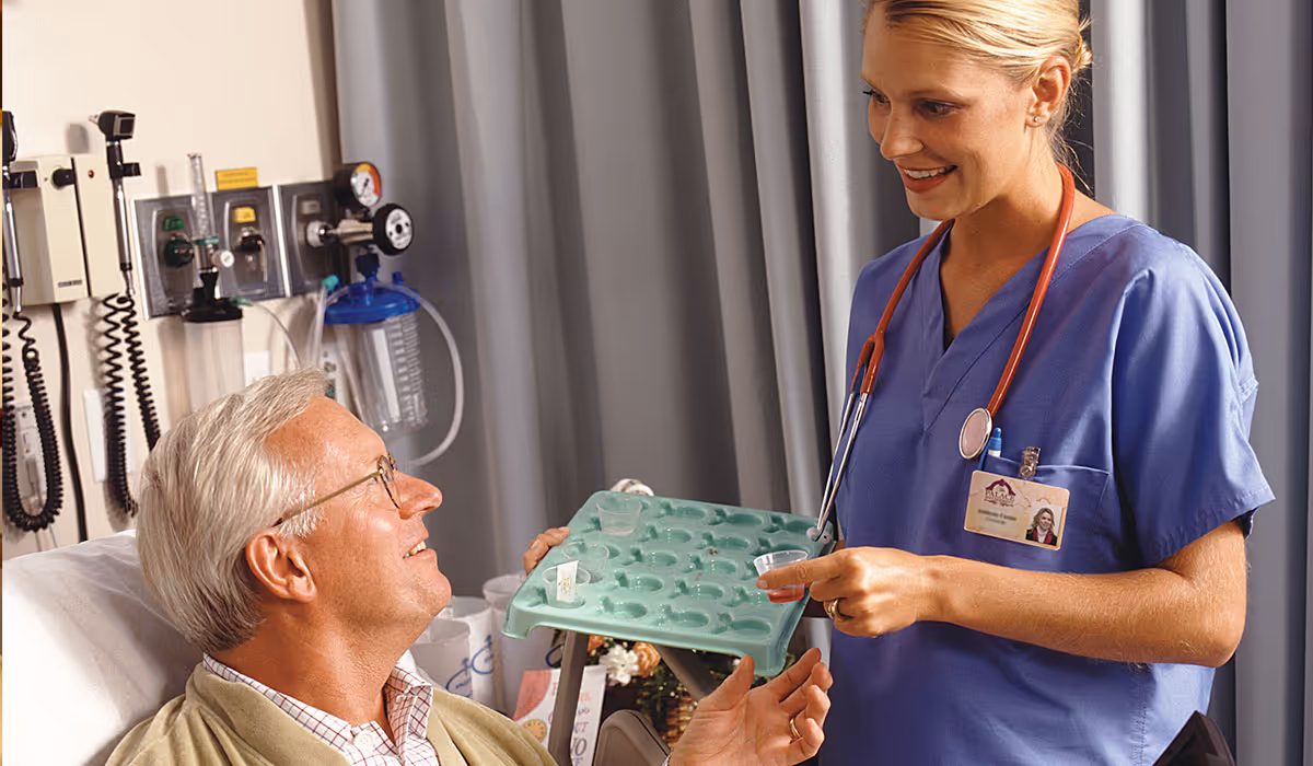 A healthcare professional in blue scrubs with a stethoscope around her neck is smiling and offering medication to an elderly male patient sitting in a hospital bed. Medical equipment is visible on the wall behind the patient.