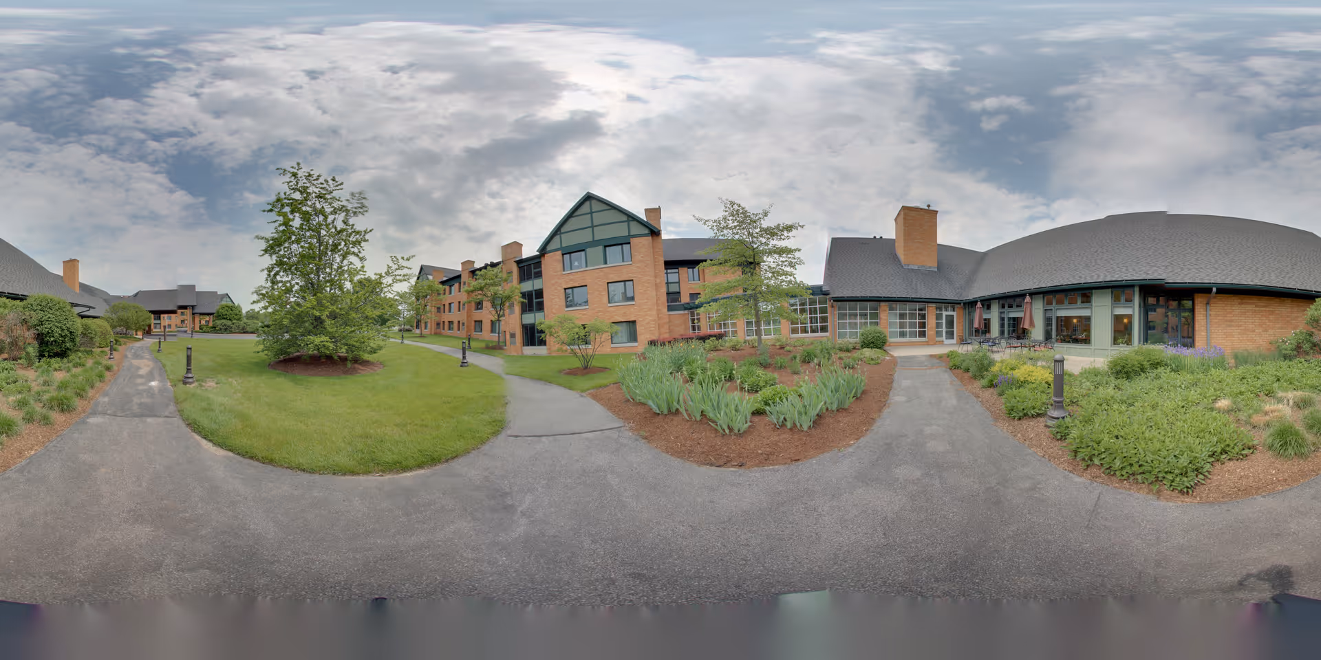 Brick senior living buildings surround a landscaped courtyard with walkways and planted beds under a cloudy sky.