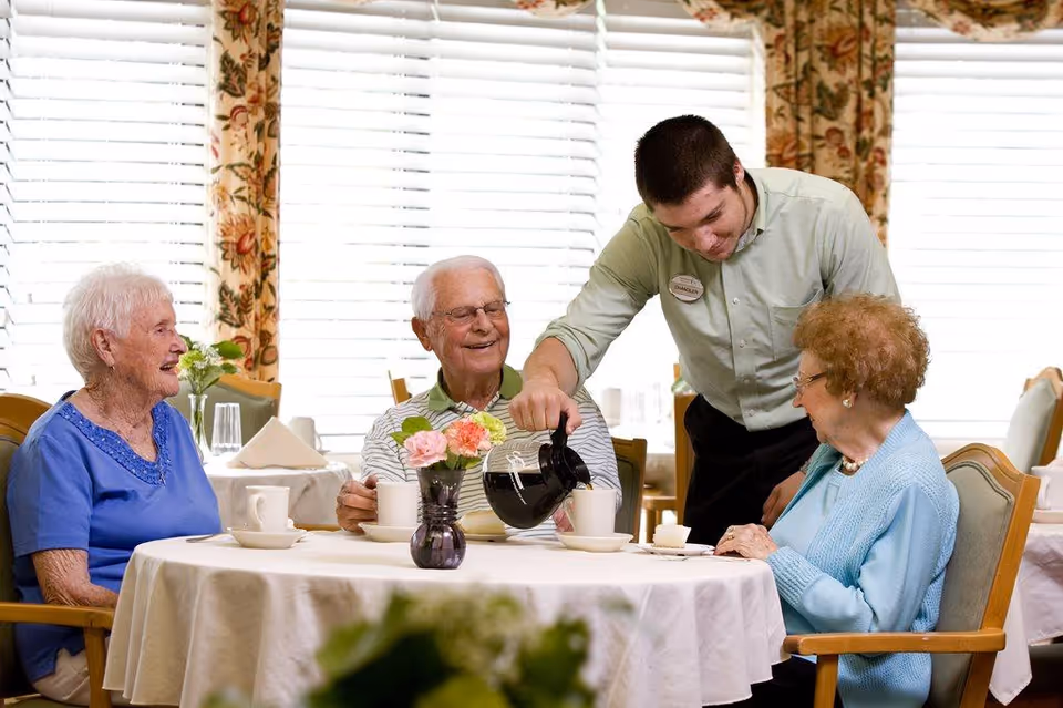 A young male staff member pours coffee for three elderly people seated around a table in a bright dining room with floral curtains. The elderly individuals are smiling and appear to be enjoying their time together.