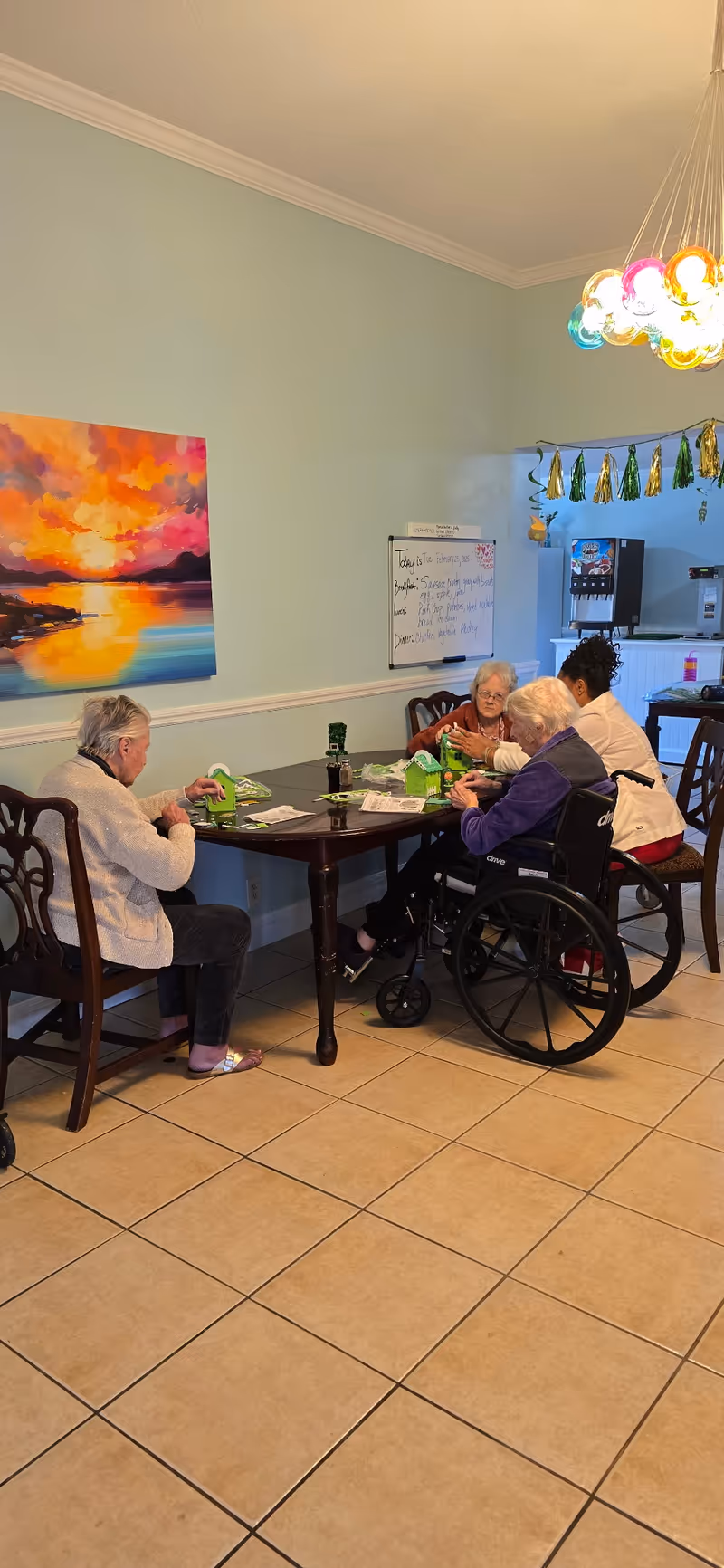 Three elderly women and a caregiver sitting around a wooden table in a well-lit room with light blue walls. Two of the elderly women are engaged in a craft activity with green materials, one seated in a wheelchair. A colorful sunset painting hangs on the wall, and a whiteboard with daily meal information is visible in the background. A colorful hanging light fixture is above the table.