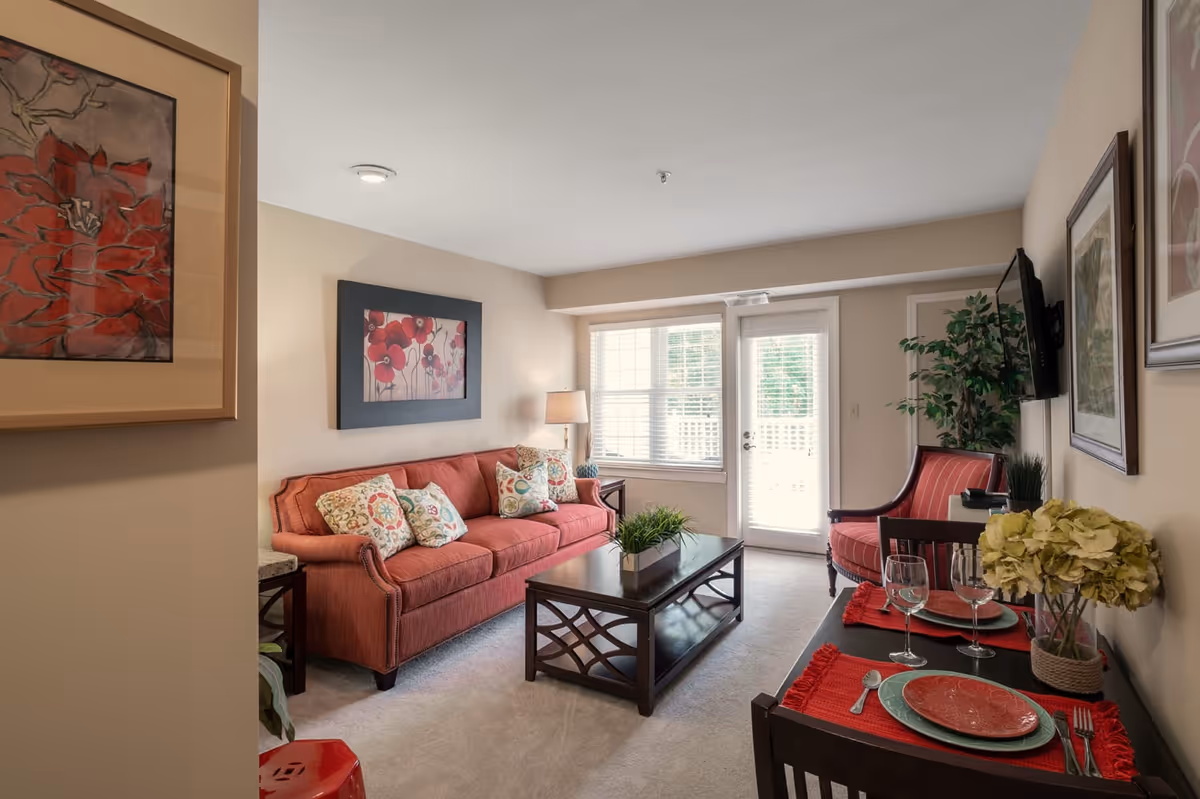 A cozy living room with a red sofa adorned with patterned cushions, a wooden coffee table with a green plant centerpiece, a red armchair, and a small dining table set for two with red placemats, plates, and wine glasses. The room has beige walls decorated with floral artwork, a window and a glass door letting in natural light, and a mounted flat-screen TV.