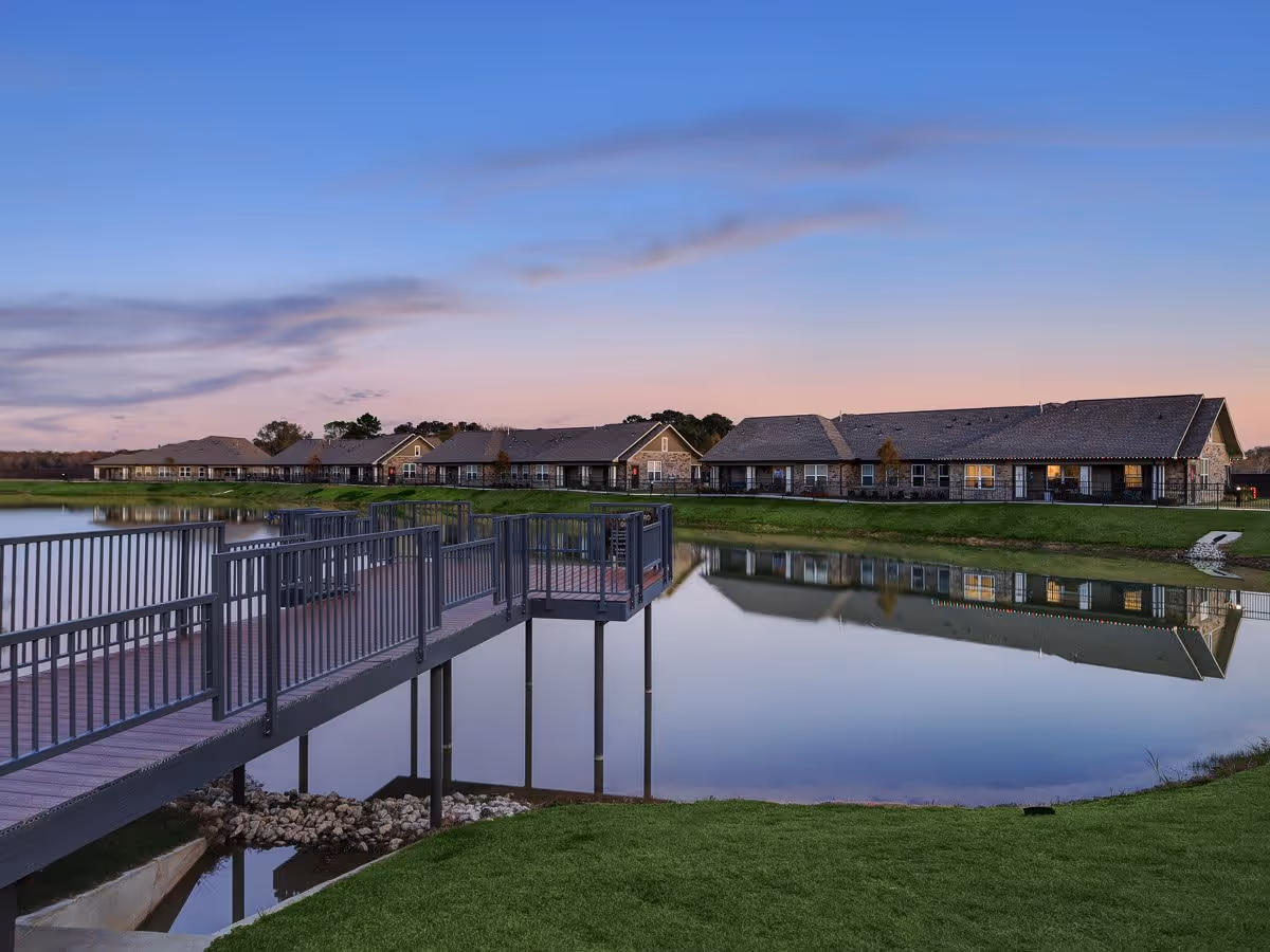 A serene outdoor scene at dusk showing a calm pond with a wooden pier extending over the water. In the background, there are single-story buildings with lit windows, reflecting on the pond's surface. The sky is clear with soft clouds and a gradient from blue to pink near the horizon.