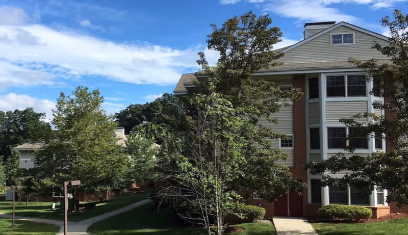 Exterior view of a multi-story residential building surrounded by trees and greenery under a partly cloudy blue sky.