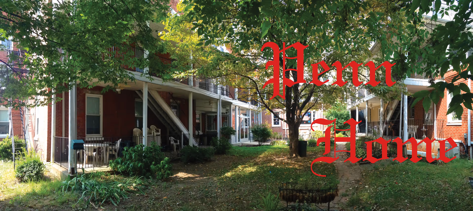 Outdoor view of a residential facility with a grassy courtyard, trees, and two-story brick buildings with balconies and stairs. There are chairs and plants on the patios and a pathway leading through the grass.