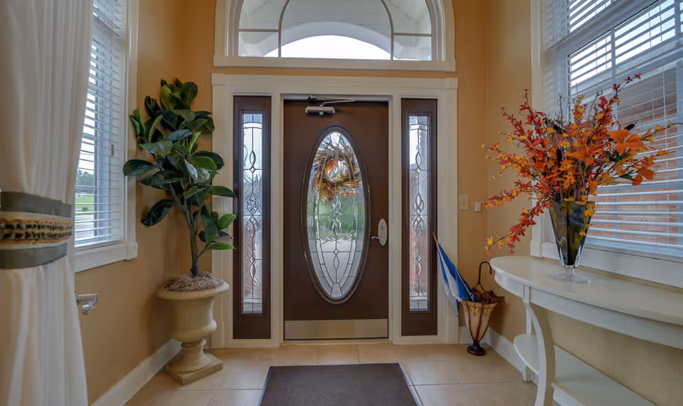 Entryway of a senior living facility with a decorative glass front door flanked by two narrow windows. There is a potted plant on the left side and a white console table with a vase of autumn-colored flowers on the right. An umbrella stand with umbrellas is next to the table. The walls are painted beige, and there are windows with white blinds on both sides.