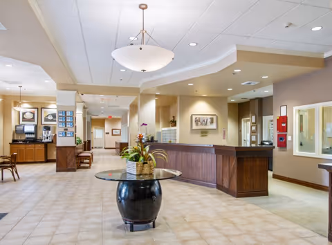 A spacious and well-lit senior living facility lobby with a round glass table holding a plant in the center. To the right is a wooden reception desk with framed pictures on the wall behind it. On the left side, there is a seating area with chairs and a coffee station. The floor is tiled, and the ceiling has recessed lighting and a hanging light fixture.