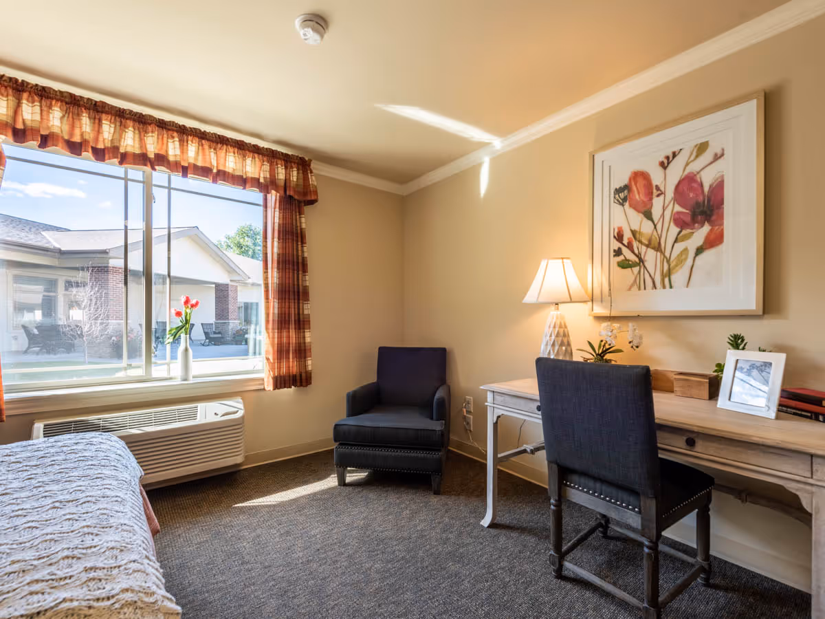 A cozy room with a large window covered by plaid curtains, a single bed with a textured blanket, a dark upholstered armchair, and a wooden desk with a chair. On the desk are a table lamp, framed photo, small plants, and decorative items. A floral painting hangs on the wall above the desk.