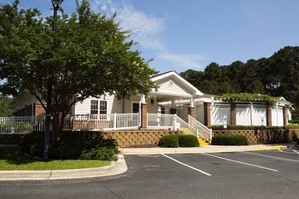 Exterior view of a single-story building with white siding and brick accents, surrounded by greenery and a parking lot with marked spaces. The building has a covered entrance with white railings and steps leading up to it, under a clear blue sky.