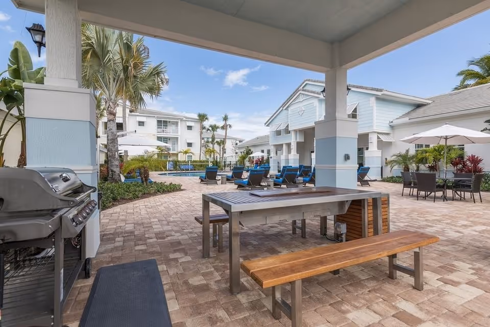 Covered outdoor patio with a picnic table and bench, barbecue grill, poolside lounge chairs and umbrellas in a residential courtyard.