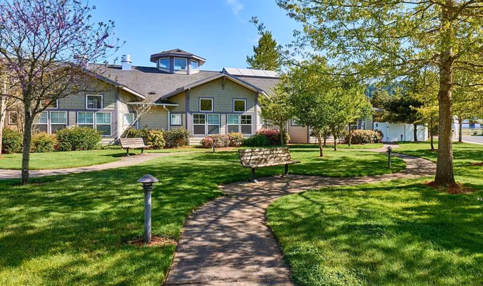 Well-maintained assisted living building front with a curved walkway, benches, green lawn, and trees.