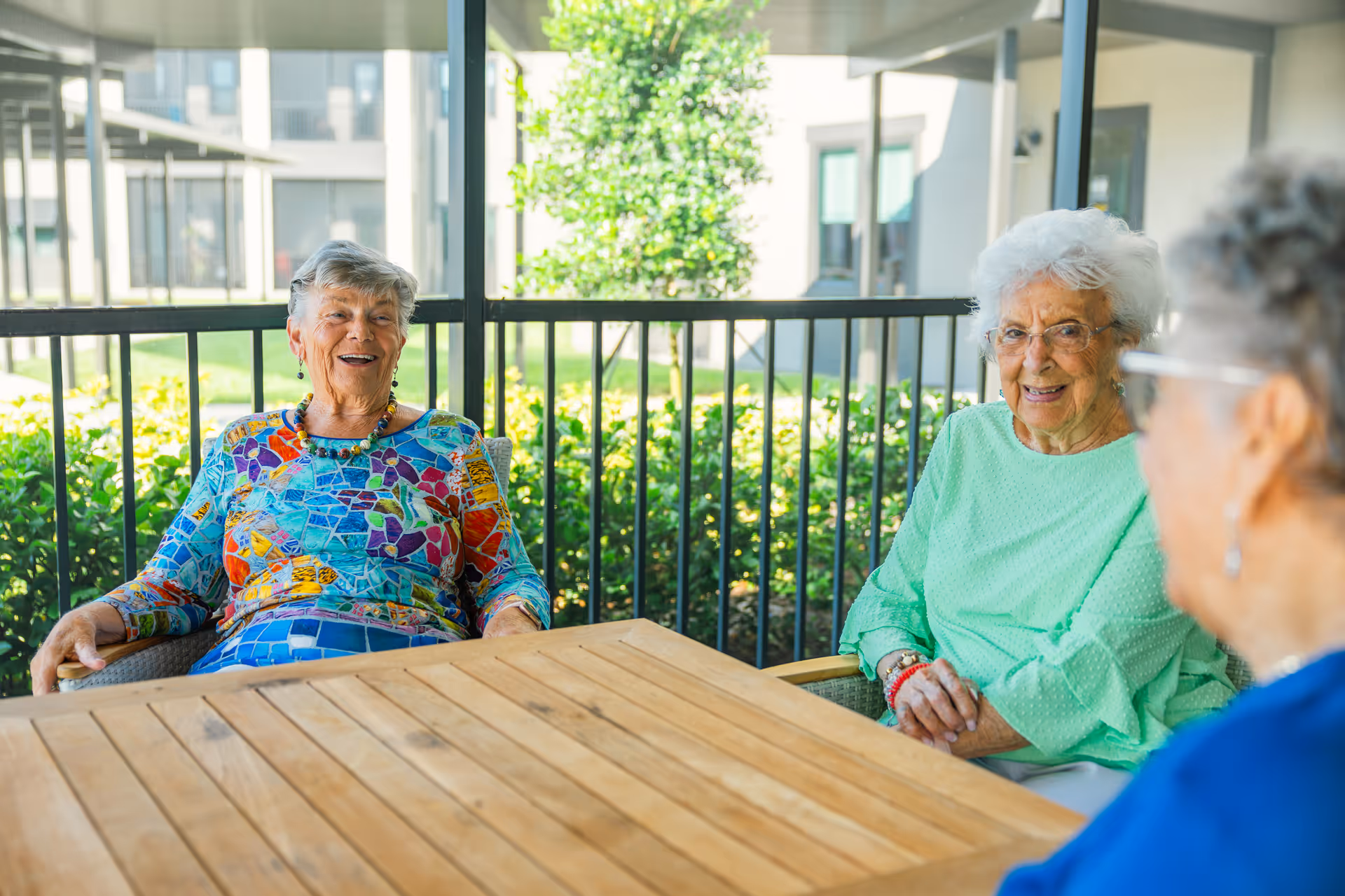 Three elderly women sitting around a wooden table on a screened outdoor patio, smiling and enjoying each other's company with greenery and building windows in the background.