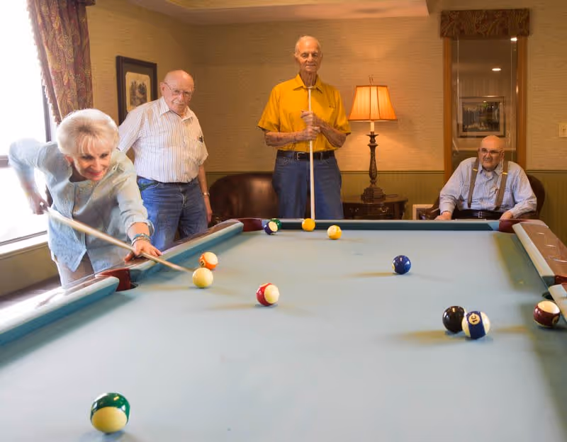 Four elderly people playing pool in a cozy room with warm lighting. One woman is taking a shot while three men watch, two standing and one sitting. The room has a lamp on a side table, framed pictures on the walls, and a window with curtains.