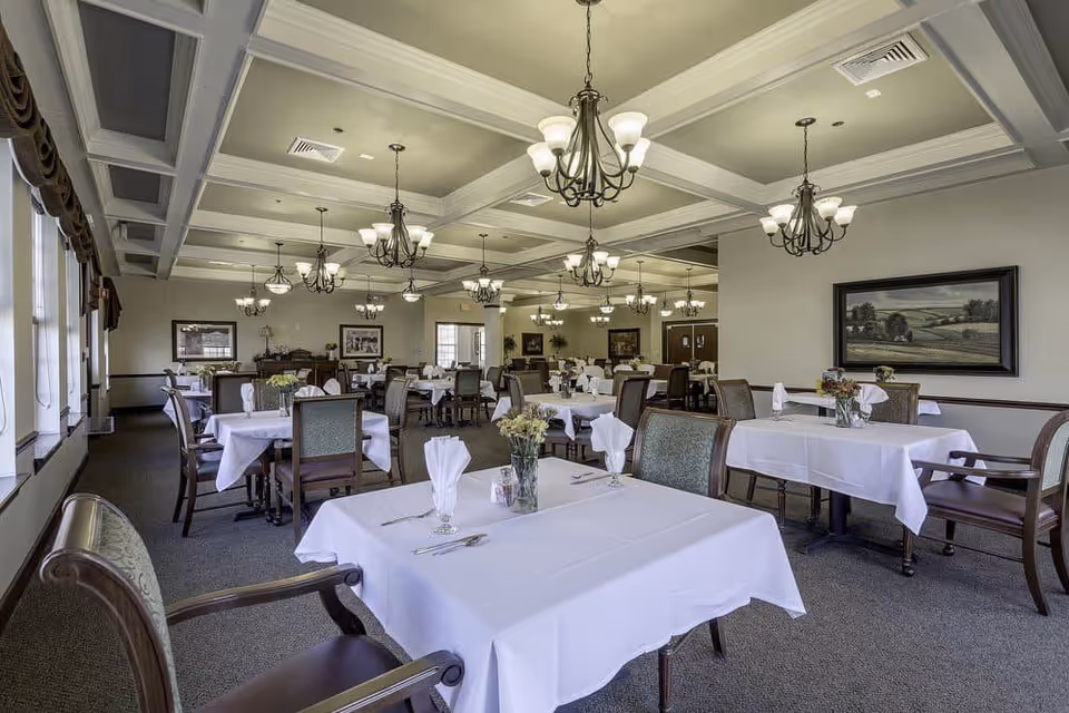 A spacious dining room with elegantly set tables, featuring white tablecloths and floral centerpieces. The room has a coffered ceiling and is well-lit with chandeliers, creating a warm and inviting atmosphere.