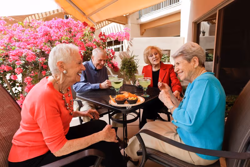 Four elderly people sitting around a black metal table outdoors, enjoying drinks and snacks. They are smiling and engaged in conversation. Behind them are vibrant pink flowering bushes and part of a building with balconies and an awning overhead.