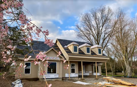 Front exterior of a cottage-style senior living building under construction with a covered porch, dormer windows, and flowering branches in the foreground.