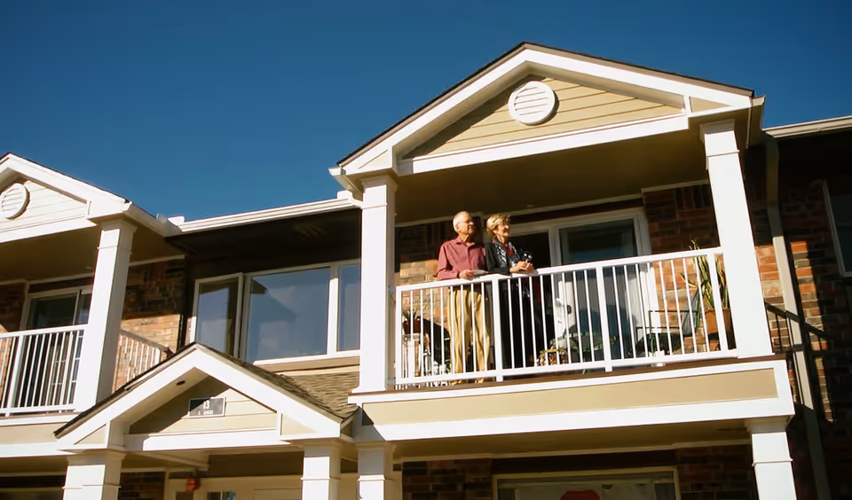 An elderly couple standing on the balcony of a two-story brick and siding building under a clear blue sky.