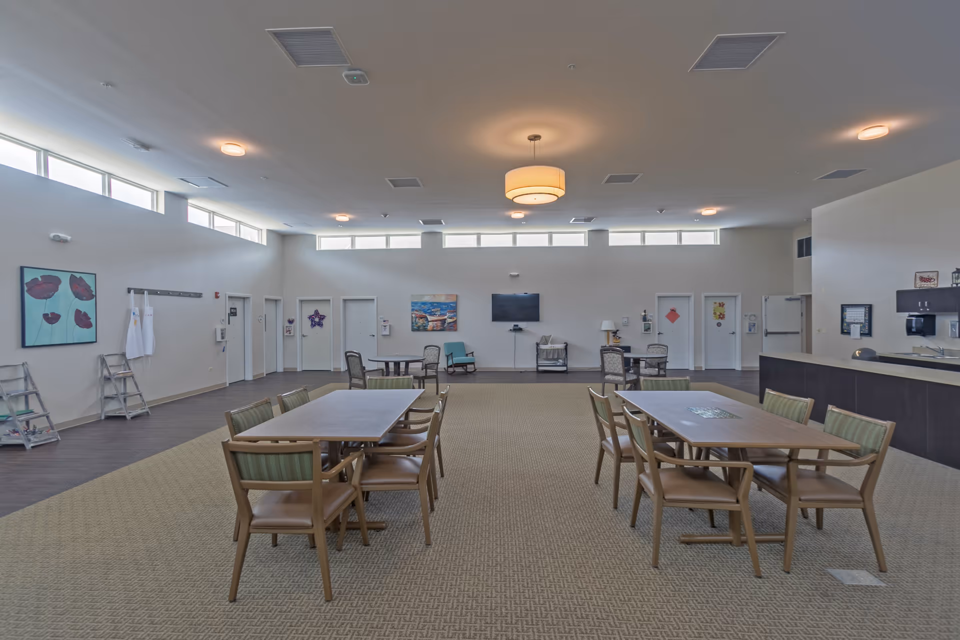A spacious common area with two rectangular tables each surrounded by six chairs. The room has high ceilings with multiple ceiling lights and windows near the ceiling letting in natural light. There is a television mounted on the far wall with a few chairs and small tables arranged around the room. The walls are decorated with paintings and there is a counter area on the right side with cabinets and a sink.