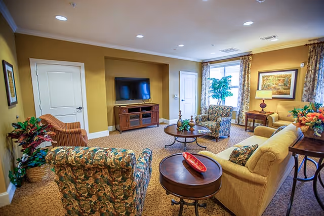 A cozy living room in an assisted living facility featuring a beige sofa, two floral-patterned armchairs, a wooden coffee table with decorative items, a TV mounted on a wooden cabinet, a side table with a lamp, framed artwork on the walls, and a large window with floral curtains letting in natural light.