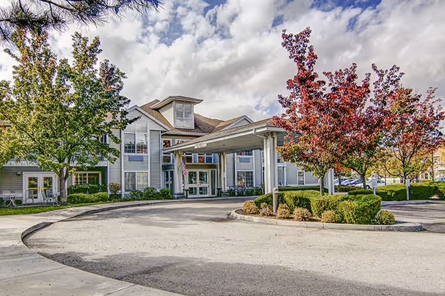 Exterior view of a senior living facility with a covered entrance, surrounded by trees with green and red leaves, under a partly cloudy sky.