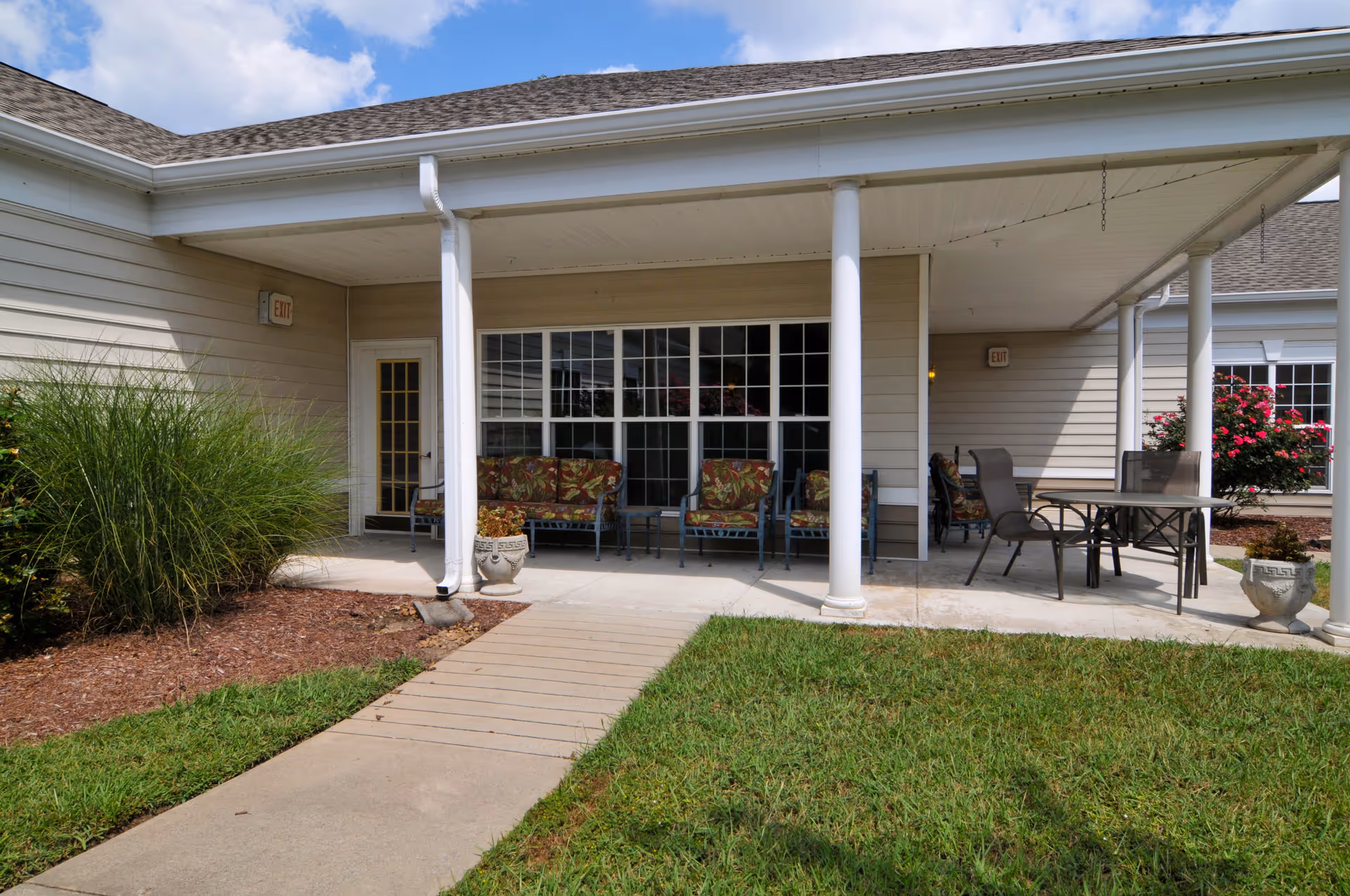 Outdoor covered patio area at Charter Senior Living of Franklin with cushioned chairs, a table with chairs, potted plants, and a walkway leading to the entrance. The building exterior is beige with white columns and large windows.