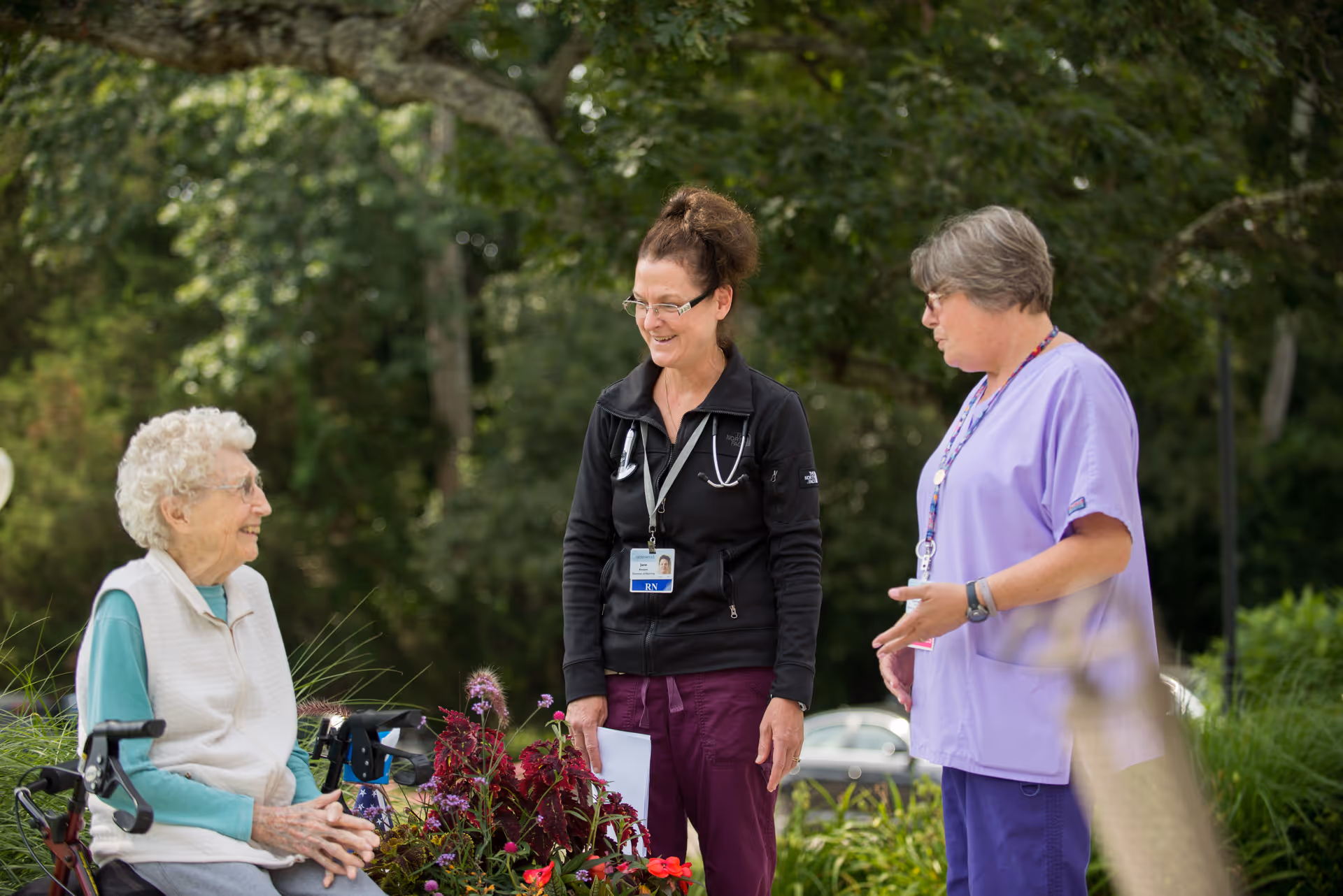 An elderly woman sitting in a wheelchair outdoors, smiling and talking with two female healthcare workers standing nearby, surrounded by greenery and flowers.