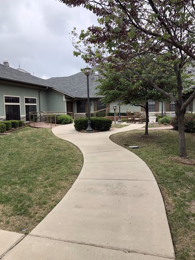 Curved concrete walkway leading through a grassy courtyard with trees and bushes, outdoor benches, and a building with green walls and multiple windows in the background under a cloudy sky.