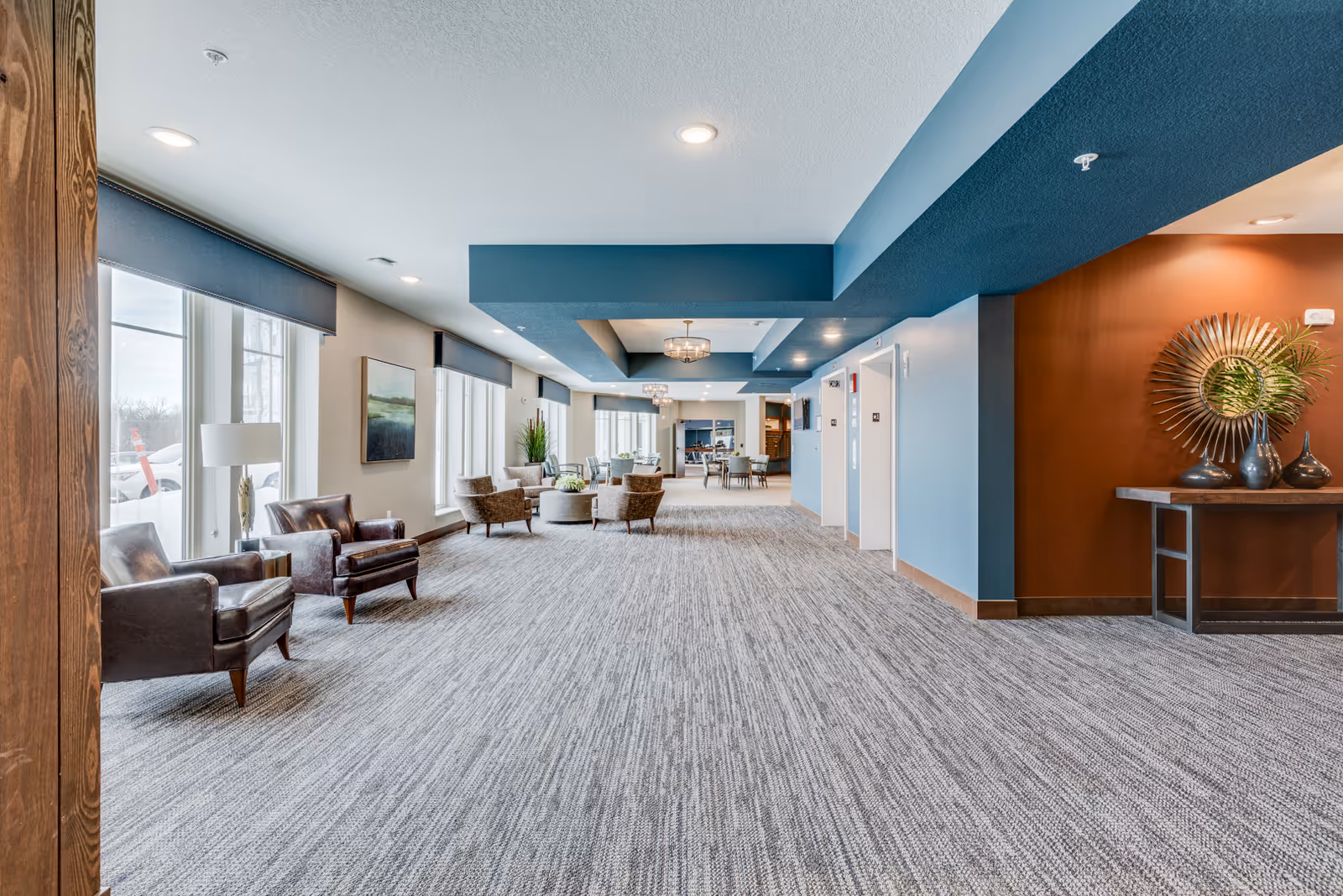 A spacious, well-lit common area in a senior living facility with large windows on the left side, several comfortable chairs arranged in small groups, modern decor including a table with decorative vases and a sunburst mirror on the right, and a carpeted floor. The ceiling features recessed lighting and a blue accent section.