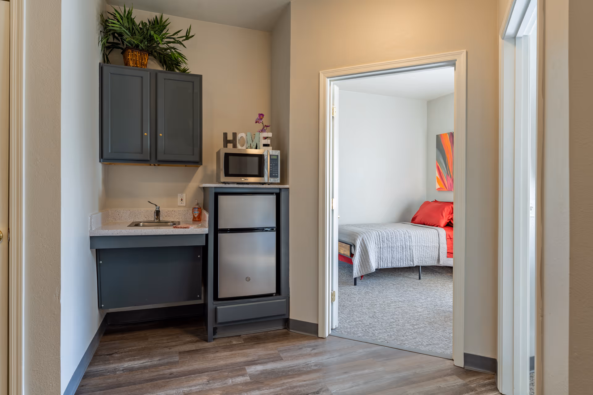 A small kitchenette area with a sink, countertop, upper cabinets, a mini refrigerator, and a microwave. There is a decorative plant on top of the cabinets and a decorative 'HOME' sign next to the microwave. Through an open doorway, a bedroom is visible with a bed covered in a gray quilt and red pillows, along with a colorful abstract painting on the wall.