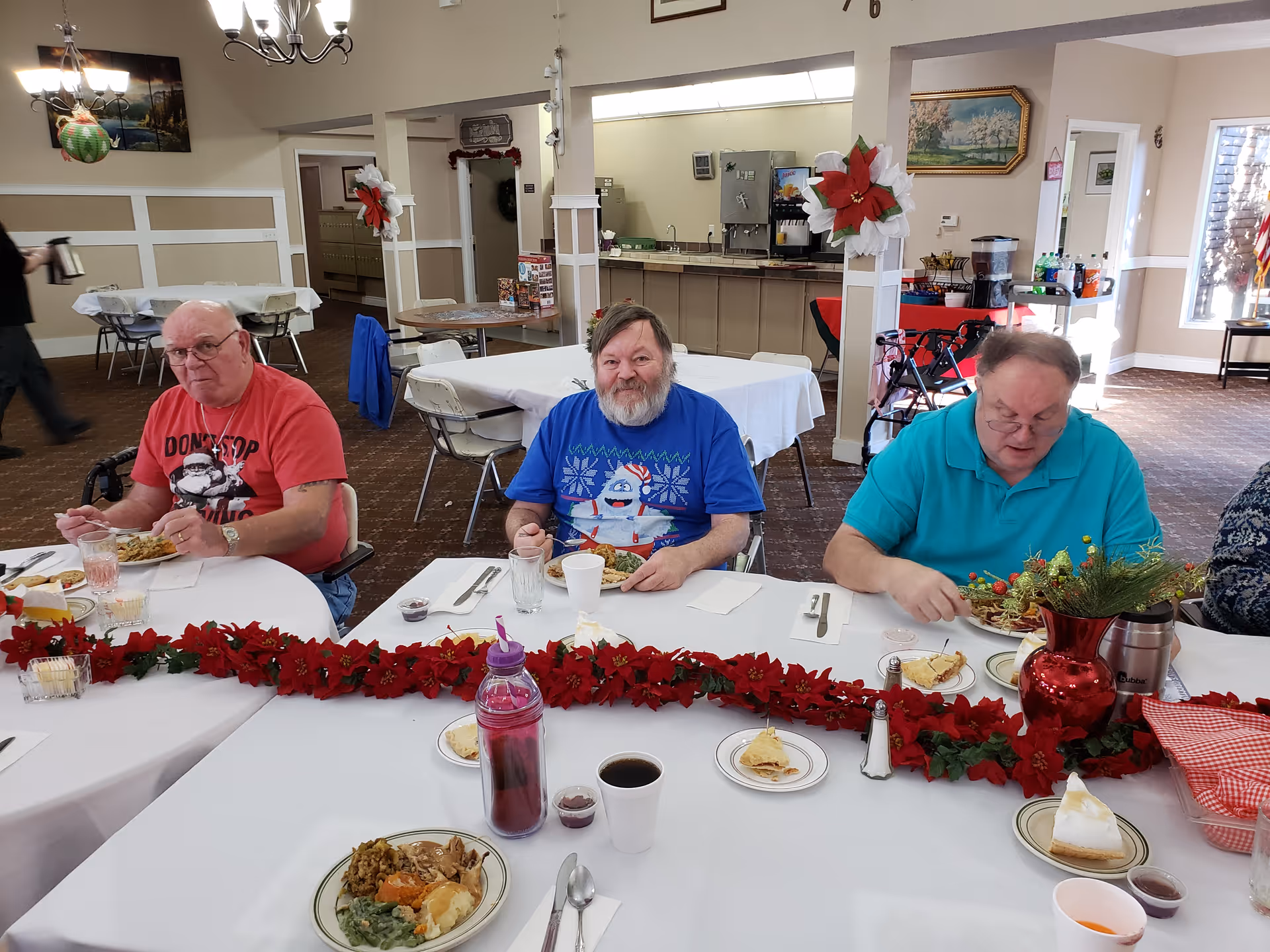 Three elderly men sitting at a decorated dining table in a retirement center, eating a meal. The table is adorned with a red garland and a red vase with greenery. The room has beige walls, white trim, and several tables and chairs. There are holiday decorations on the walls and columns.