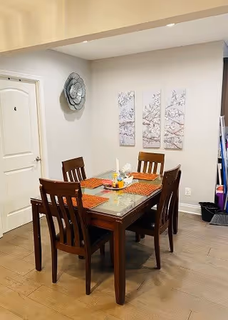 A dining area with a wooden table and four matching chairs. The table has a glass top and is set with orange placemats and a centerpiece. On the wall behind the table, there are three decorative panels featuring a floral design and a large metallic flower decoration. A closed white door is visible to the left, and some shelves with items are partially visible on the right.