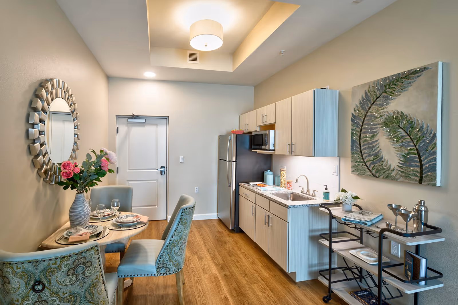 A small, modern kitchen and dining area with light wood cabinets, a stainless steel refrigerator, a microwave, and a granite countertop with a sink. A round dining table set for four with patterned upholstered chairs and a vase of pink flowers is on the left. A decorative round mirror hangs on the wall above the table, and a large artwork featuring green leaves is on the wall above a small rolling cart with shelves holding books, glasses, and a cocktail shaker. The room has wood flooring and soft lighting from a ceiling fixture.