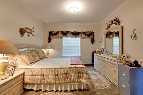 A cozy bedroom with a large bed covered in beige and floral bedding. There are two bedside tables with lamps on either side of the bed. A window with a decorative valance is centered on the far wall. On the right side of the room, there is a long dresser with a large mirror above it, decorated with various personal items and flowers. The walls are light-colored, and the room has a warm, inviting atmosphere.