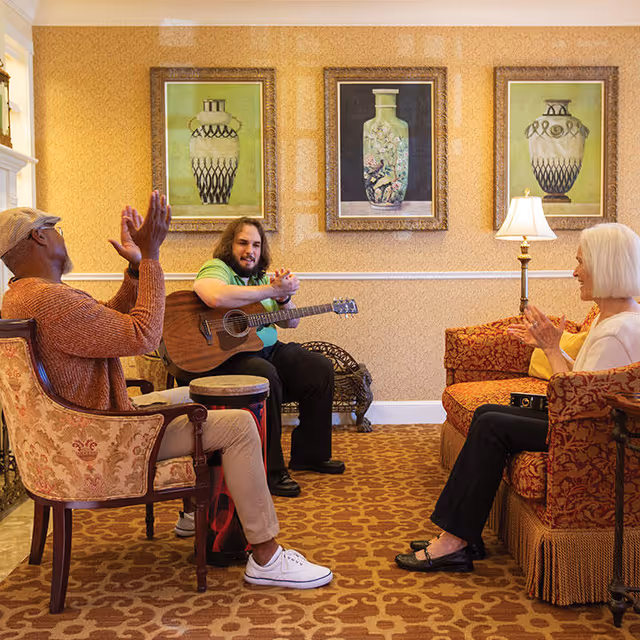 Three people in a cozy living room setting at Parc Provence. One person is playing an acoustic guitar while the other two, seated on upholstered chairs and a sofa, are clapping along. The room features patterned carpet, framed artwork of vases on the wall, and a floor lamp providing warm lighting.