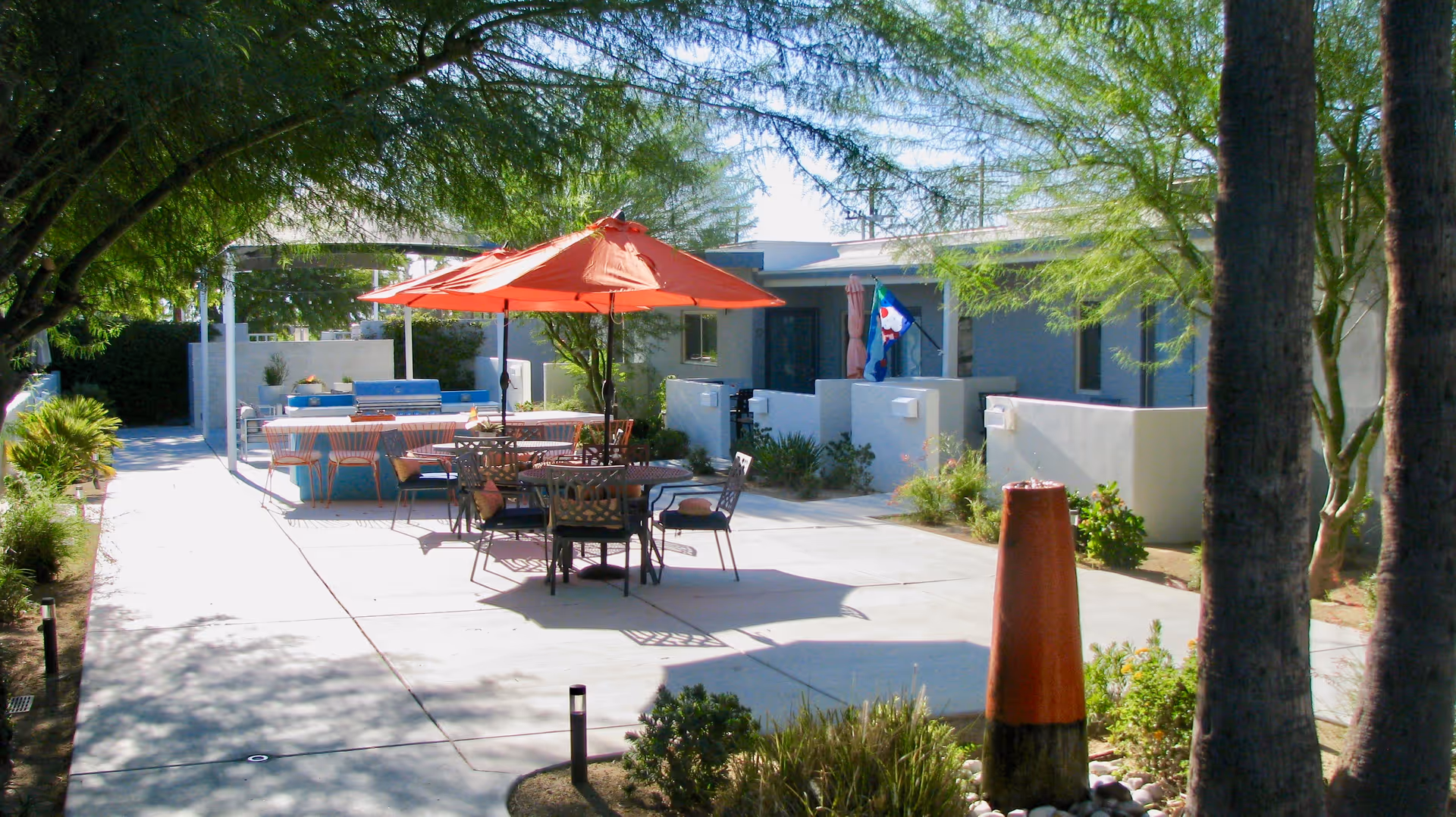 Outdoor patio area at Stonewall Gardens Assisted Living with tables and chairs under a large orange umbrella, surrounded by trees and plants, with a pathway leading through the space.