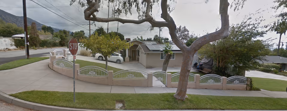 Exterior view of a single-story residential building with a beige facade, solar panels on the roof, and a small front yard enclosed by a low wall with decorative metal fencing. A large tree is in the foreground, and a stop sign is visible on the sidewalk near the street corner.