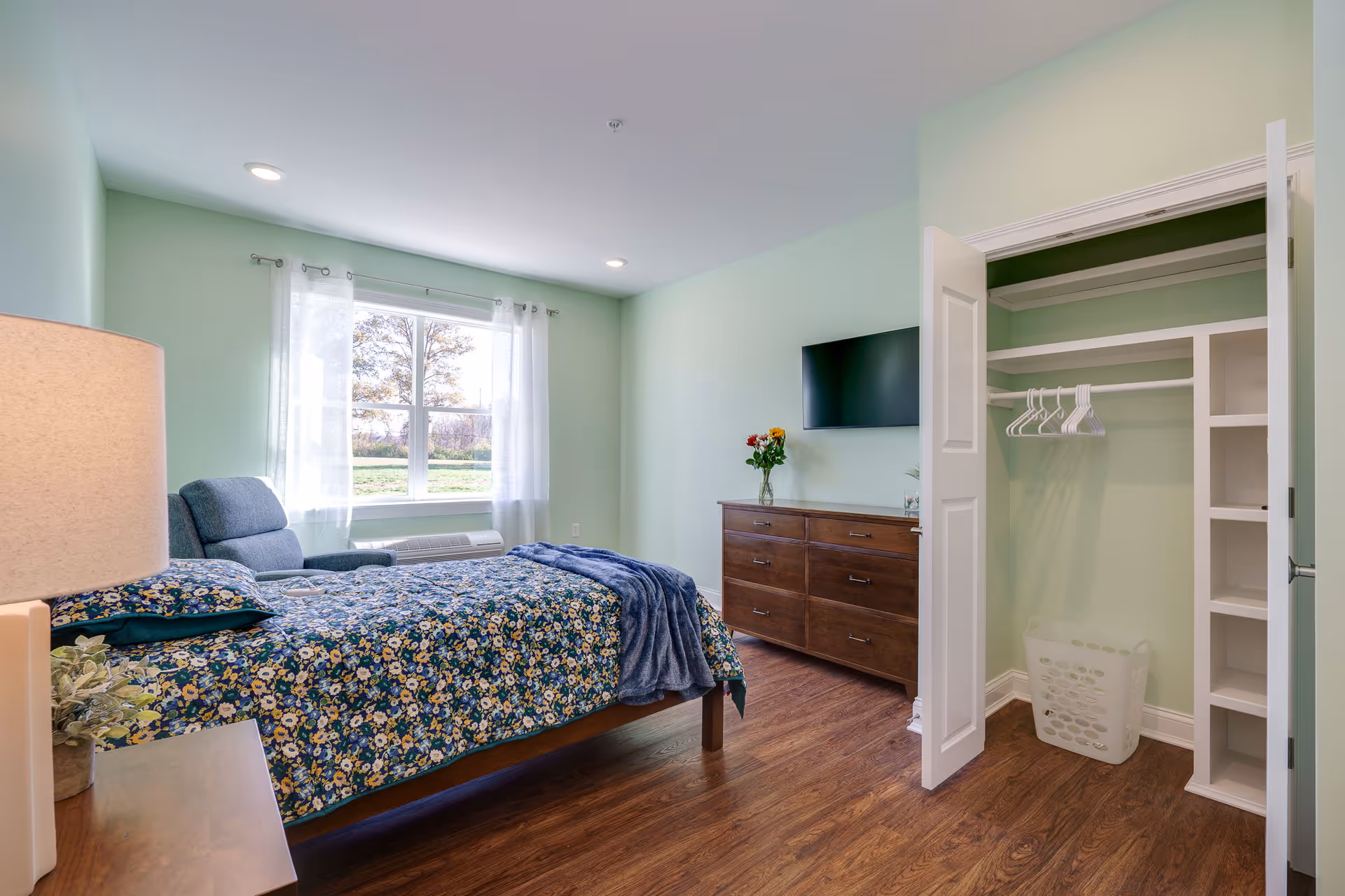A bright bedroom with light green walls and wood flooring. The room features a bed with a floral bedspread and a blue throw blanket, a wooden dresser with a vase of flowers, a wall-mounted flat-screen TV, an open closet with white hangers and shelves, and a window with sheer white curtains letting in natural light.